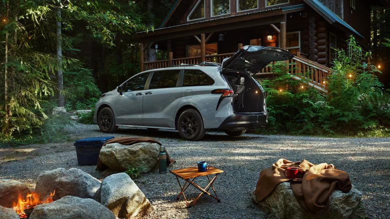 A white Toyota Sienna rear three-quarters view of the left side with its tailgate open outside a woodland cabin.