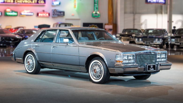 A pale blue Cadillac Seville from between 1976 to 1979 parked inside a large garage with other cars out of focus in the back.