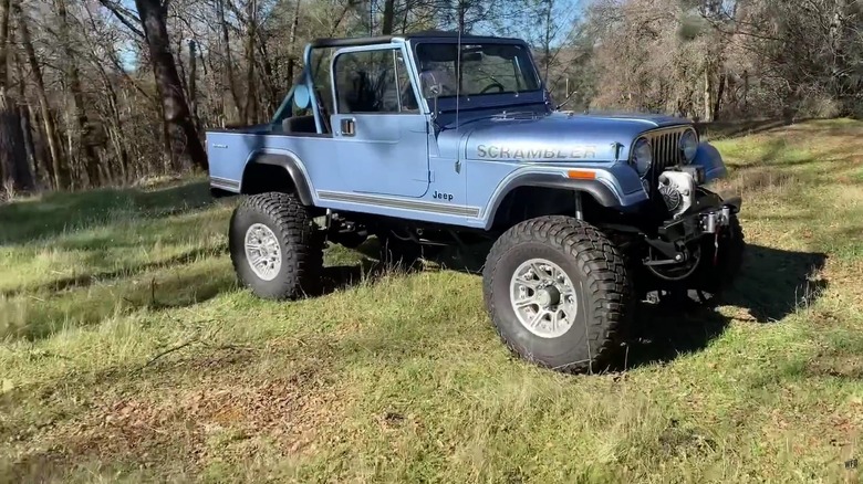 A pale blue Jeep Scrambler CJ-8 pickup truck from the 1980s standing in a forest in the afternoon.