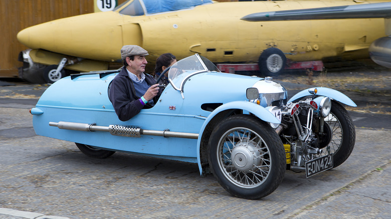 Sky blue Morgan 3 Wheeler in front of yellow jet fuselage
