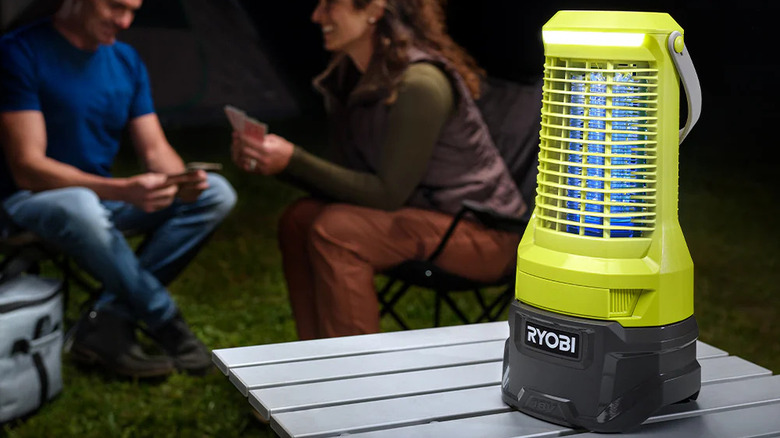 Ryobi's Bug Zapper on a table at a campsite, two people sit in the background