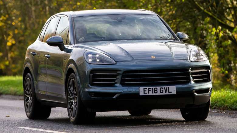 A dark green Porsche Cayenne driving down a country road with greenery in the background.
