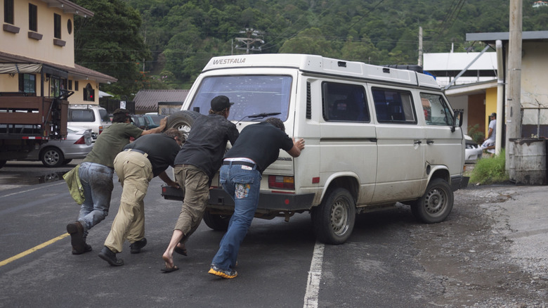 A broken down Volkswagen Vanagon being pushed by four people into a workshop.