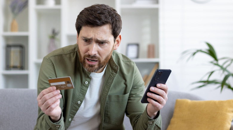 Man holding smartphone while looking at credit card with an upset expression on his face