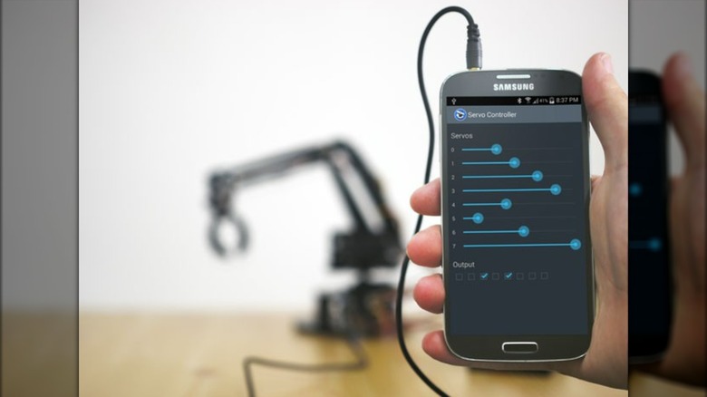 A Samsung Android smartphone showing the ServoDroid app interface with a robot arm behind it.