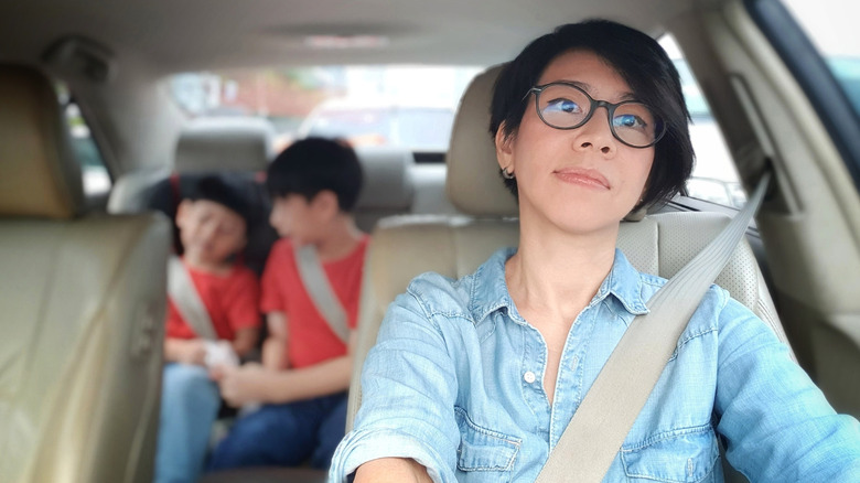 Woman with dark hair and glasses driving a car with two small children in the back seat behind her.