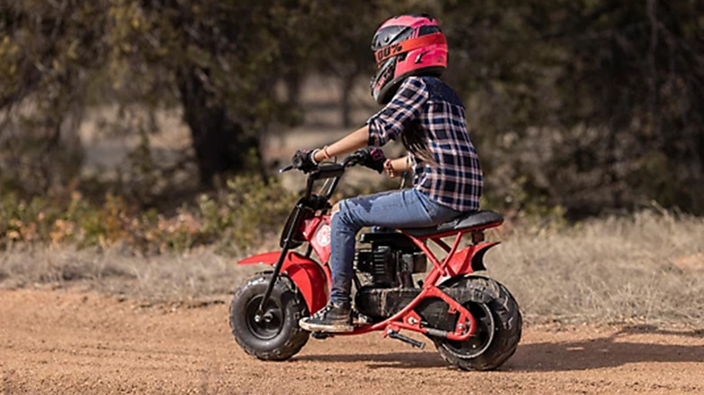 young person in red helmet, jeans, and flannel shirt riding Coleman B100 mini bike on dirt road