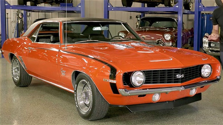 A presenter stanings next to a bright orange 1969 Camaro SS 396 inside a garage, with several other classic cars visible on lifts in the background.