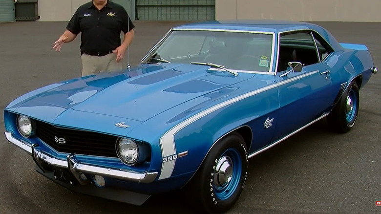 A man in a black polo shirt standing beside a brilliant blue 1969 Camaro SS 396 in an outdoor parking lot, gesturing towards the car.