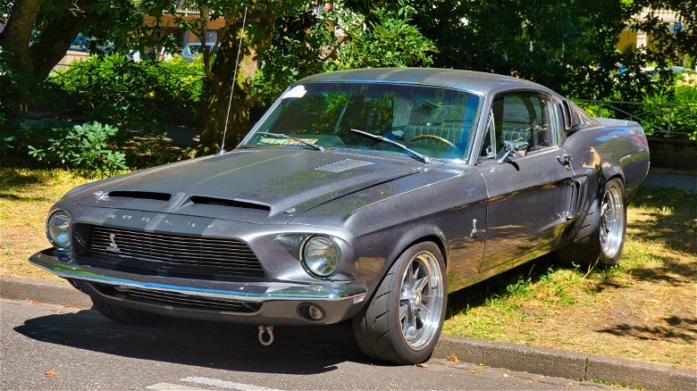 A sage green Shelby GT350 fastback sitting on a gravel lot near a chain-link fence with tall pine trees in the background.
