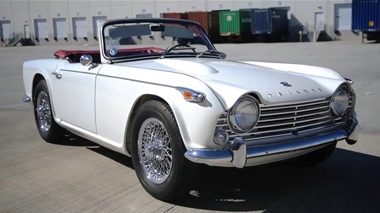 A white 1961 Triumph TR4 convertible with its top down sitting on an open concrete lot outside a shipping warehouse.