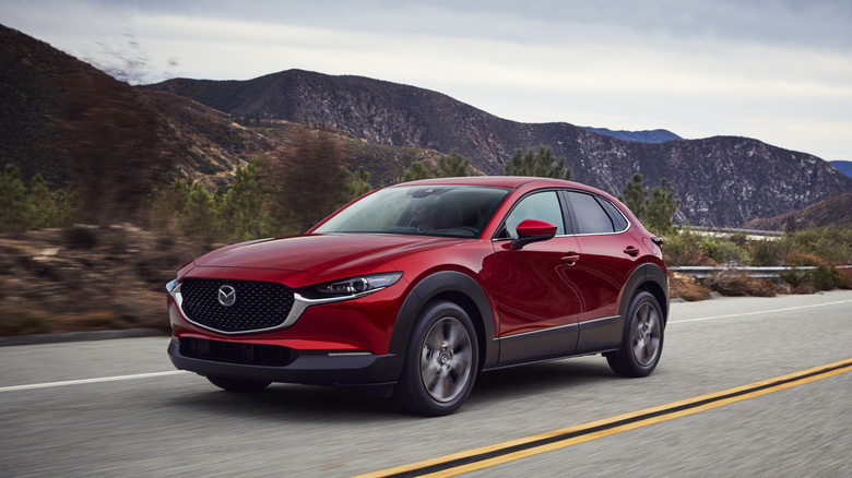 A metallic red Mazda CX-30 subcompact crossover SUV moving down a road at speed in the evening, with mountains in the back.