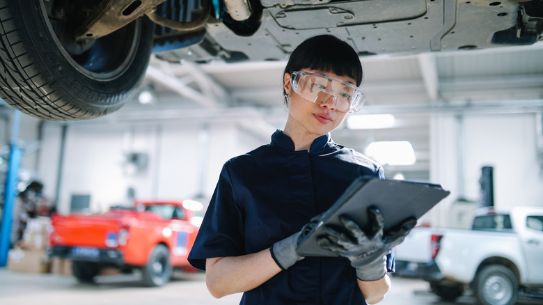 A female technician standing under a car with protective glasses while looking at a form.