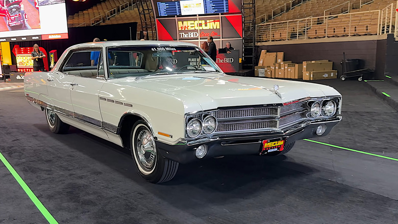A cream 1965 Buick Electra 225 hard top cruiser rolling down a passage at an indoor auction, chrome accents glinting.
