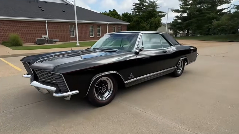 A black 1965 Buick Riviera Gran Sport coupe muscle car parked at an angle in an empty concrete lot, left three-quarters view visible.