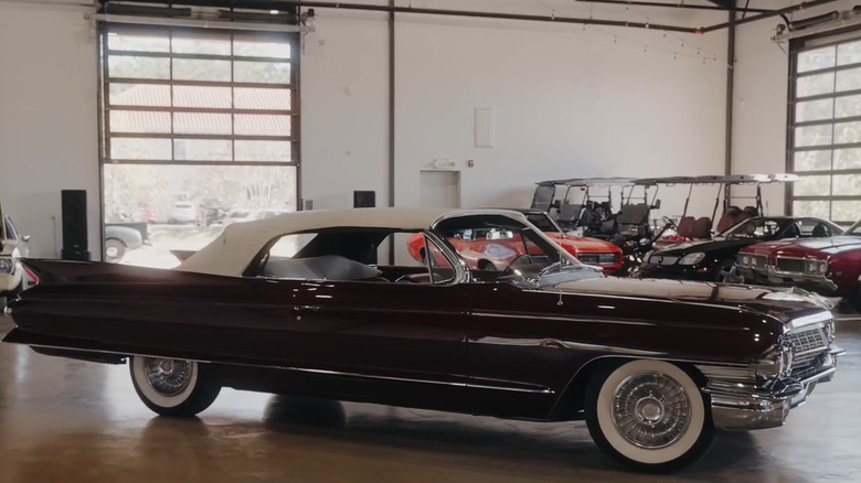 A convertible Cadillac Series 62 coupe car with whitewall tires standing inside a garage with other classic cars visible in the back.