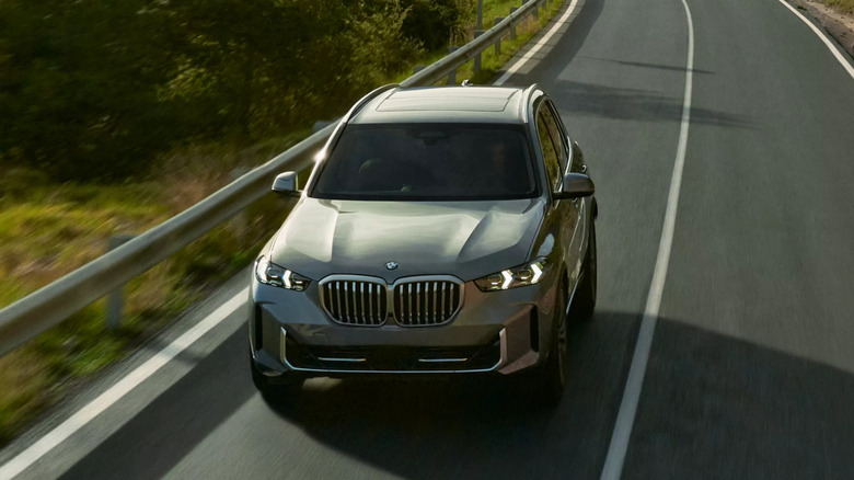 A gray BMW X5 from the 2026 model year moving at speed down a narrow asphalt road with greenery visible in the back.