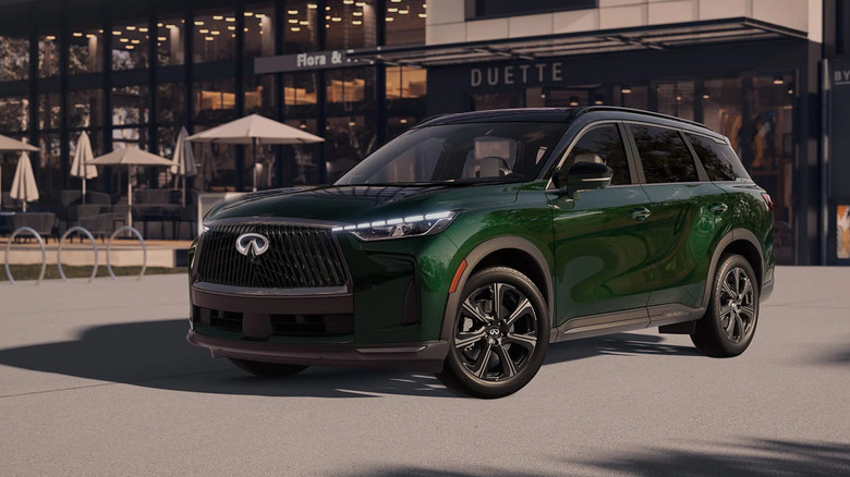 A dark green 2026 Infiniti QX60 seven-seater SUV parked outside a modern glass-fronted building with outdoor seating.