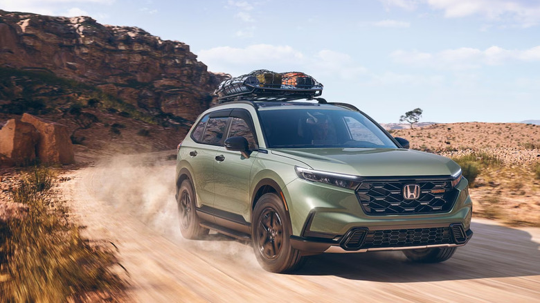 A green Honda CR-V kicking up dust as it moves along a cliffside road at speed with mountains in the backdrop.