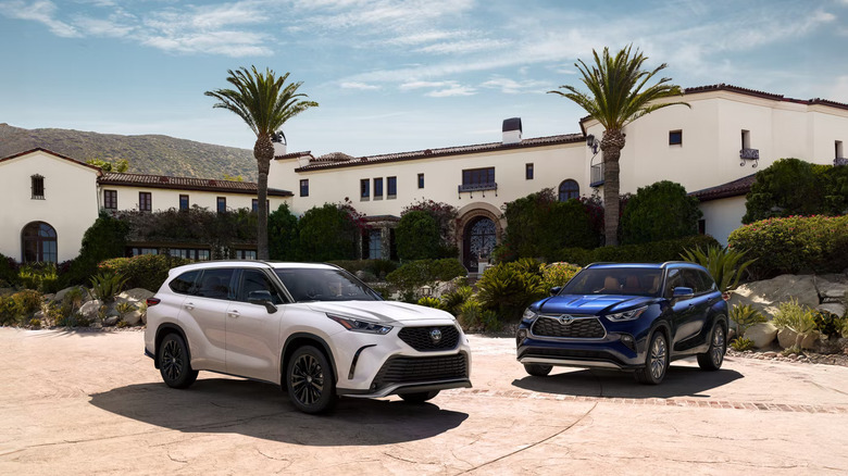 A pair of Toyota Highlander SUVs, both for the 2026 model year, one finished in white and the other in blue, parked in a suburban cul de sac.