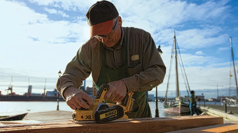 A craftsman using a DeWalt hand planer on a piece of wood with a dock background