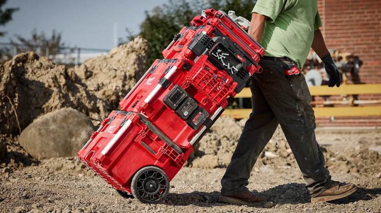 A person rolling a stack of red Milwaukee Packout boxes across dirt and rubble