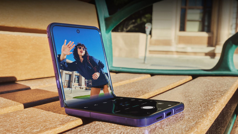 The 2026 Motorola Razr Ultra, half-folded on a wooden bench with a person waving at the camera on the screen
