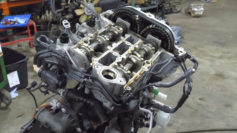 A disassembled Ford 1.0-liter EcoBoost engine sitting on a stand inside a busy workshop with its valve train, camshafts, and timing components fully exposed under the shop lights.