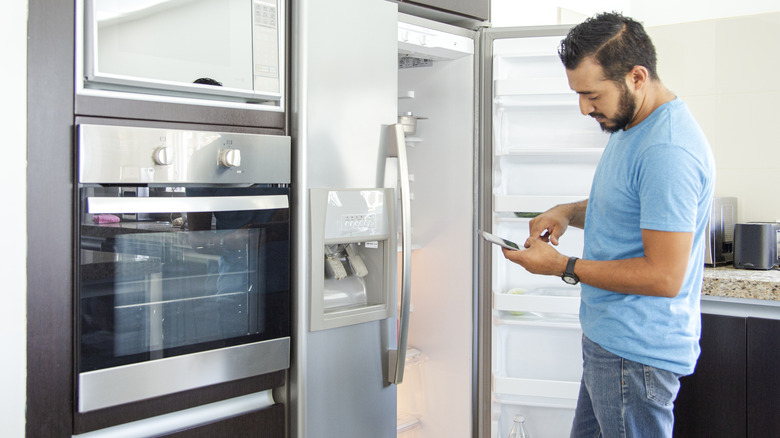 man with smartphone standing in front of open fridge