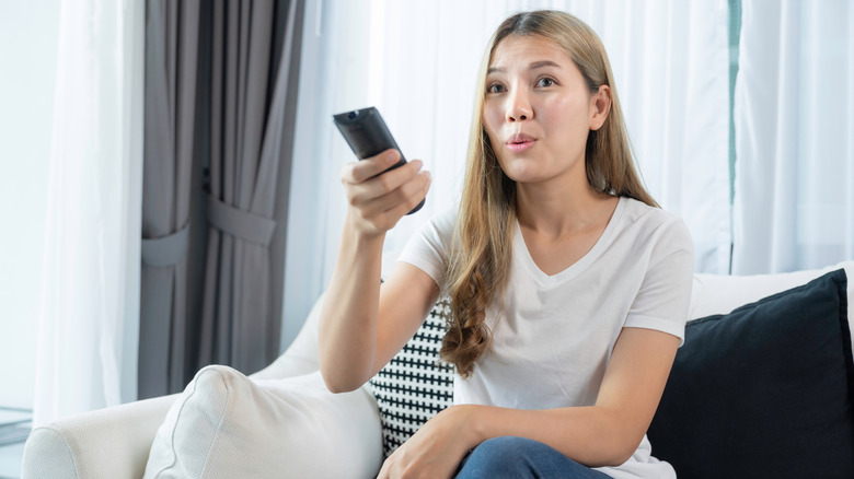 A woman points a remote to a TV behind the camera, looking confused.