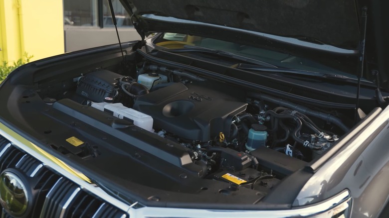 The engine bay of a modern Toyota SUV with the hood open, showing a clean, plastic-covered 1GD-FTV engine layout inside a Land Cruiser–style front end.