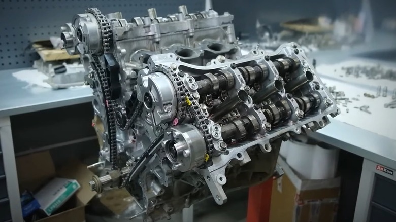 A partially disassembled aluminum 1GR-FE engine sitting on a stand in a workshop, with its camshafts, timing chain, and valve components clearly exposed.
