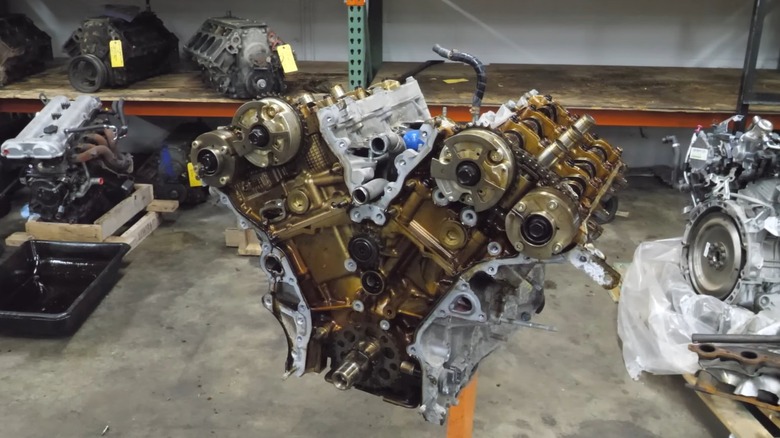 A used 2GR-FE engine mounted on a stand inside a parts warehouse, surrounded by shelves filled with other removed engines and drivetrain components.