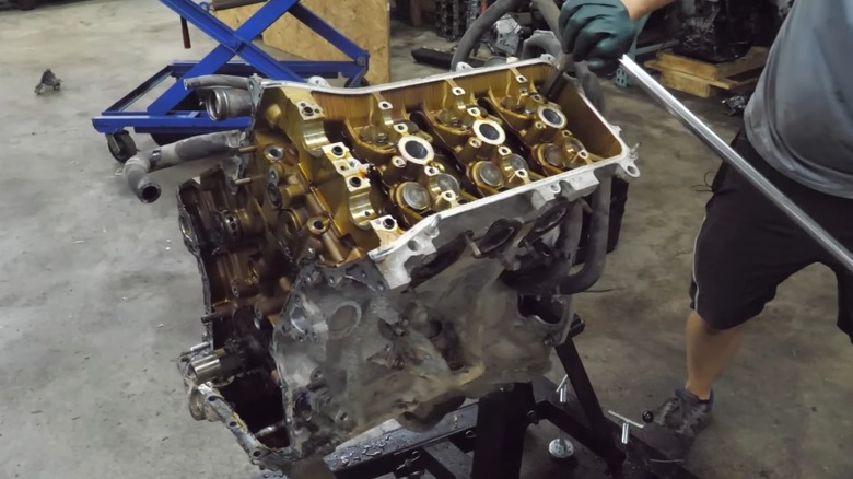 A guy in a workshop using a breaker bar to loosen parts on a partially disassembled 1GR-FE engine sitting on a stand with tools  scattered around the shop.