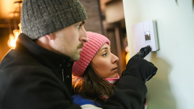 Two people wearing hats and gloves looking at a thermostat on a wall