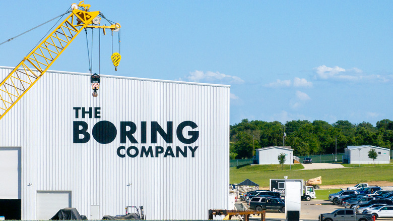 An industrial warehouse with The Boring Company logo on the front with a yellow crane visible