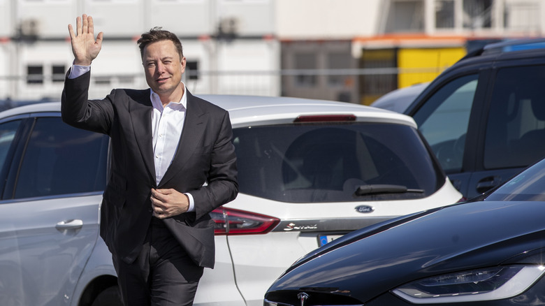 Elon Musk posing for a photograph with his hand raised and smiling