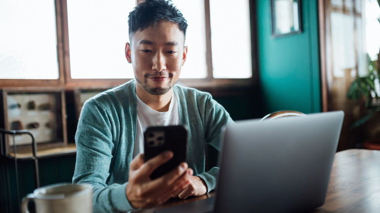 A person holding a phone while using a laptop and sitting on a wooden desk