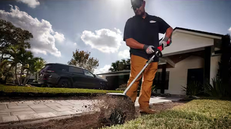 A man using a Milwaukee cultivator to dig a trench next to a driveway.
