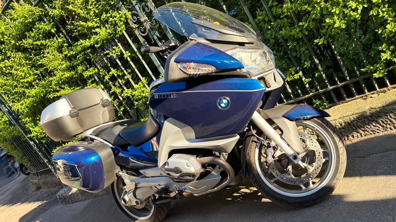A blue and silver BMW R1200RT motorbike from 2012 parked at the side of a road with foliage in the background.