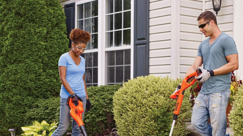 Two people using two Black and Decker, one holds a blower and the other a string trimmer.