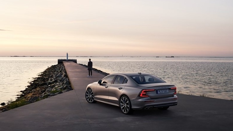 A 2021 Volvo S60 finished in pebble grey on a jetty with the sea as a background, rear and left profile of the car visible in the evening sunset