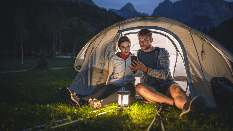 Two people sitting in front of a camping tent next to a lantern, with one of them using her phone