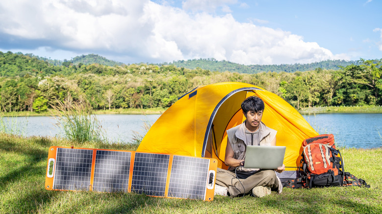 A person camping while using a laptop and connecting it to a solar charger, in front of a camping tent next to their backpack