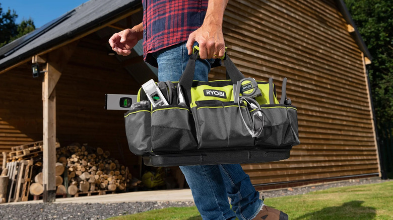 A man carrying a Ryobi tool bag through a yard near a garage.
