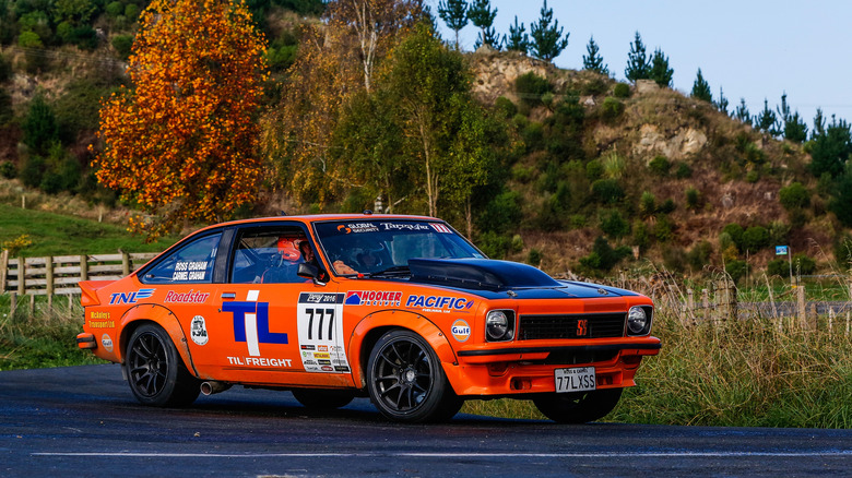 An orange 1977 Holden Torana with sponsor decals being driven as part of the Targa Rally in Australia, with foilage in the backdrop.