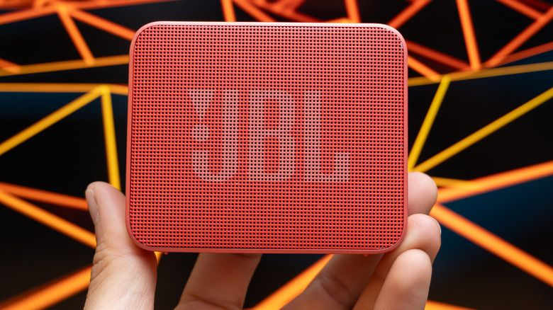 Hand holding a small JBL waterproof speaker with black and orange geometric background