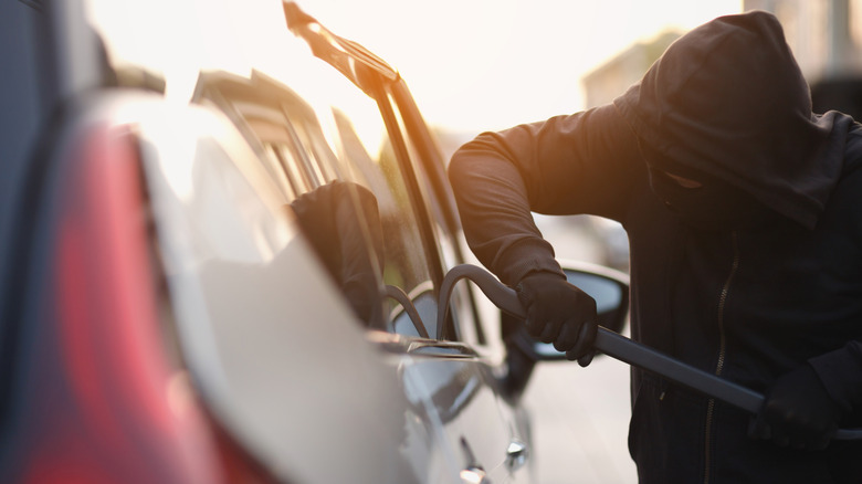 A hooded person breaking into a white vehicle with a prybar.