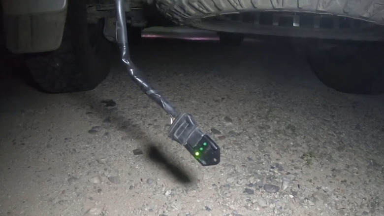 A device with one lit LED hangs from a cable under a vehicle