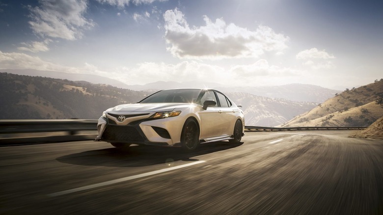 White Toyota Camry driving on a highway with hills and a cloudy blue sky in the background